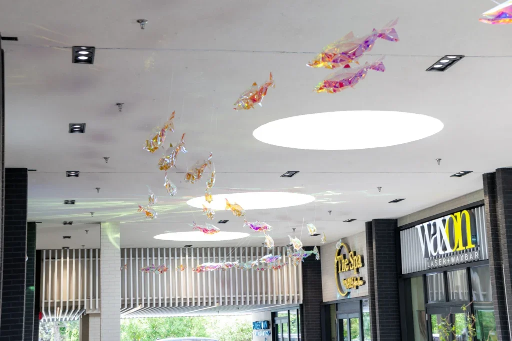 Shopping walkway with suspended fish installation overhead and a central planter, with a person walking beneath.