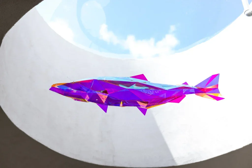 Faceted, iridescent fish sculpture suspended by thin wires beneath a circular skylight, reflecting purple and pink tones.