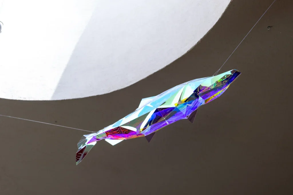 Close-up of a suspended geometric fish sculpture, refracting blue, green, and purple light against a neutral ceiling.