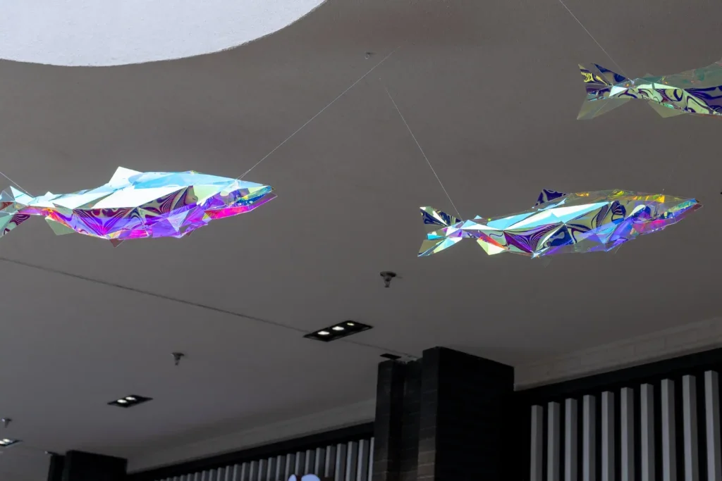 Two angular, iridescent fish sculptures hanging from the ceiling, connected by thin wires in an indoor space.