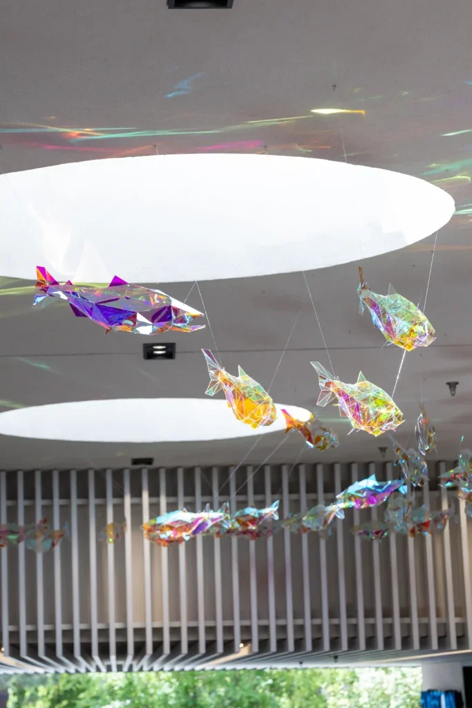 Multiple suspended fish sculptures in varying colors forming a school beneath circular ceiling skylights.