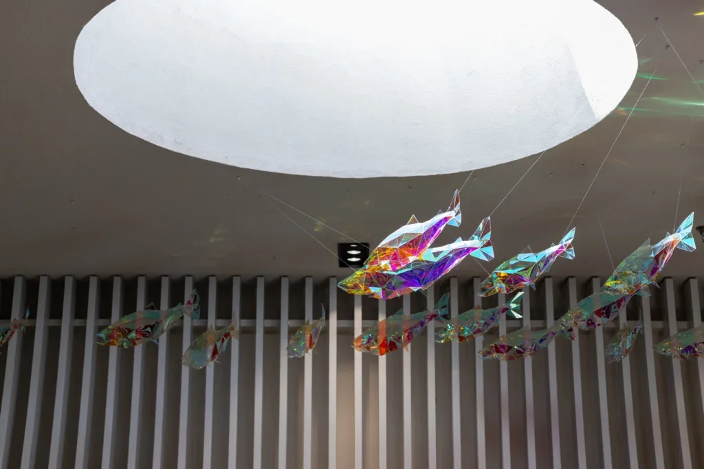 Cluster of translucent, prism-like fish sculptures hanging in a row, reflecting multicolored light.