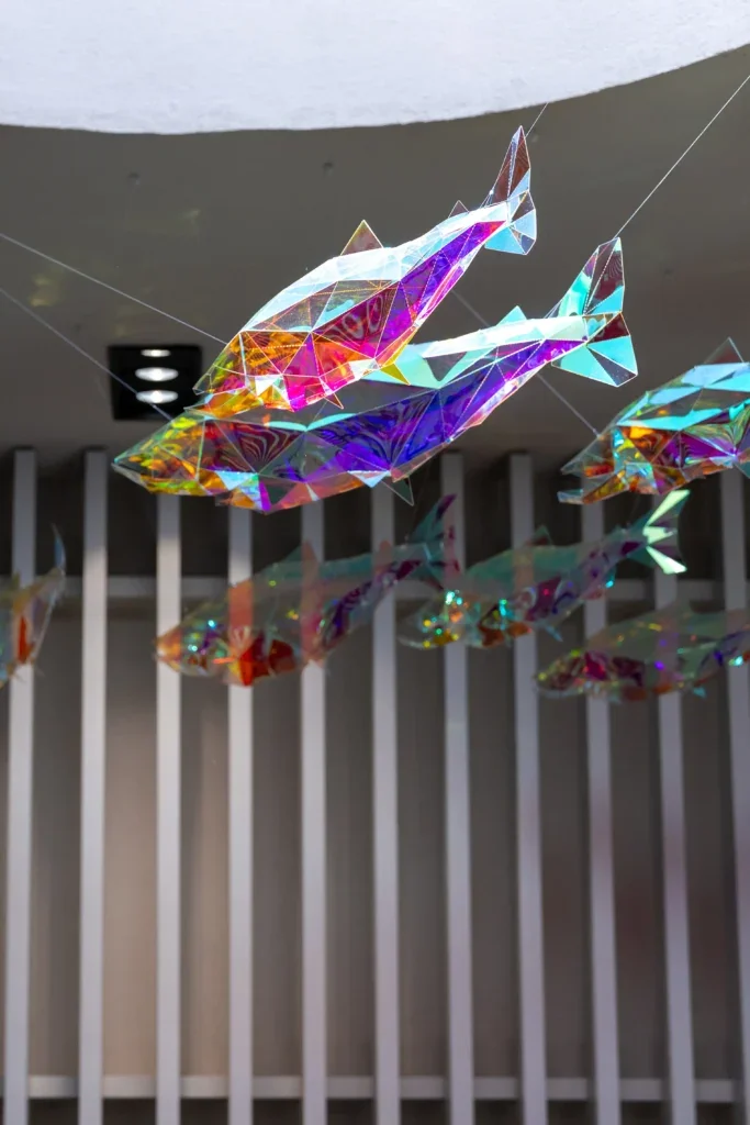 Close-up of two suspended fish sculptures with vivid rainbow reflections against vertical slatted wall panels.