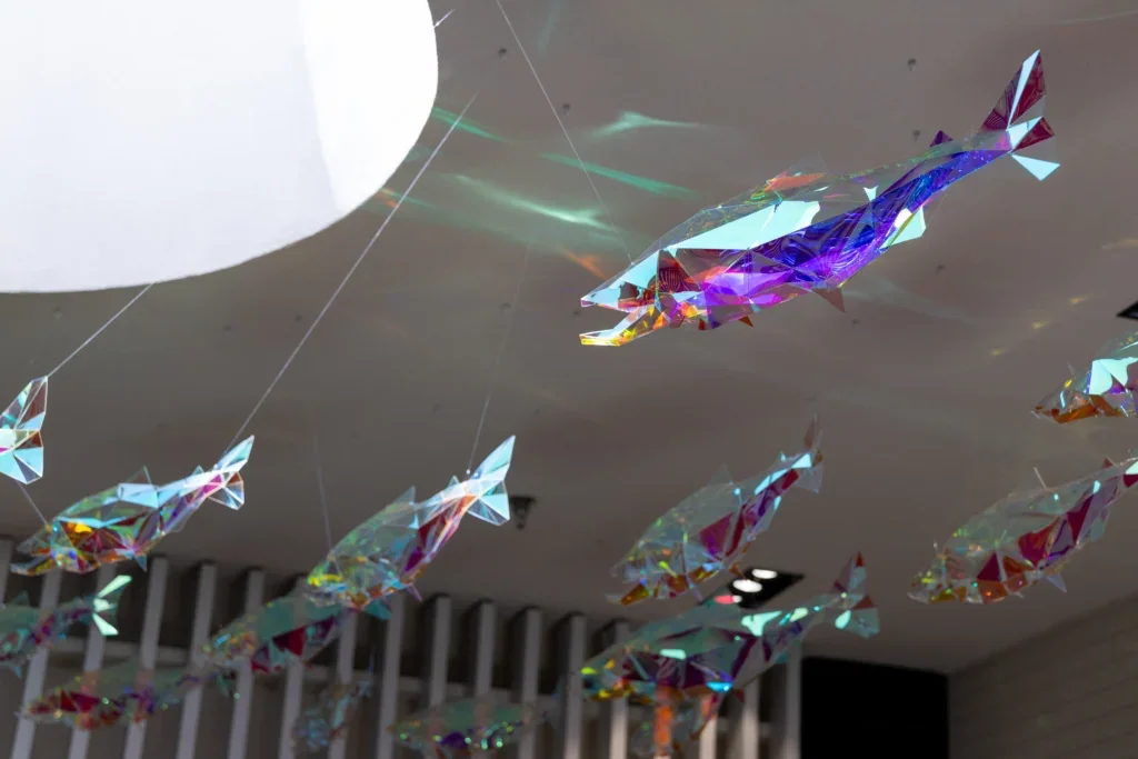 Single iridescent fish sculpture viewed from below with light reflections scattered across the ceiling.