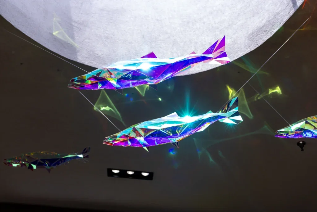Close-up of two hanging fish sculptures glowing with bright, refracted light near a circular ceiling opening.