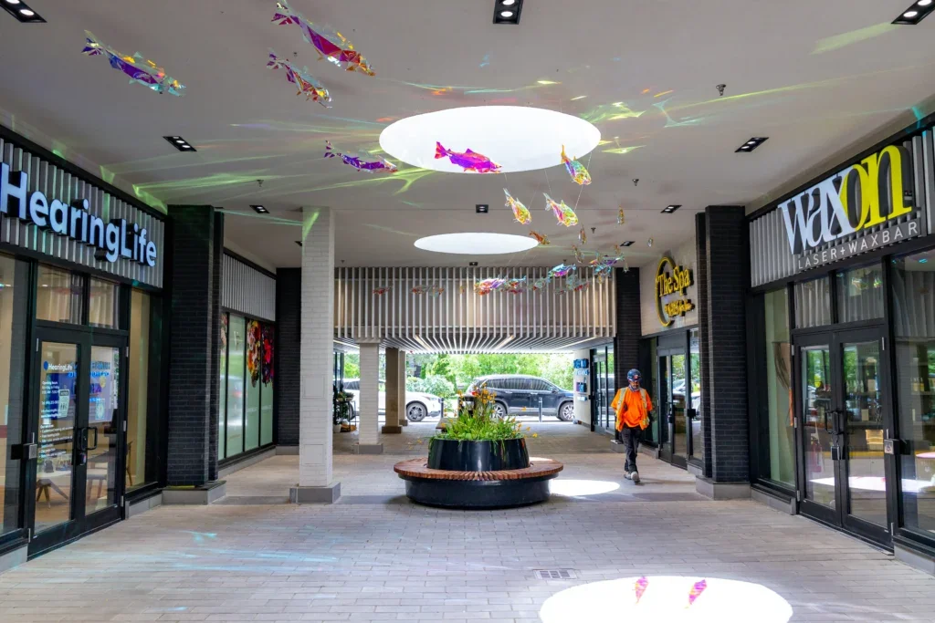 Wide shot of a corridor with multiple fish sculptures suspended overhead and storefront signs on both sides.