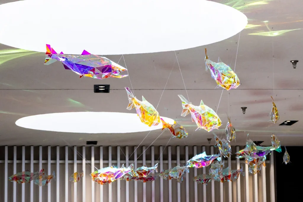 Extended view of the installation showing numerous fish sculptures hanging in a flowing arrangement across the ceiling.